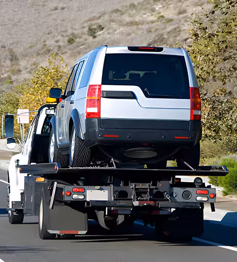 A silver suv on the back of a flatbed tow truck driving down the a surface street away from the camera.